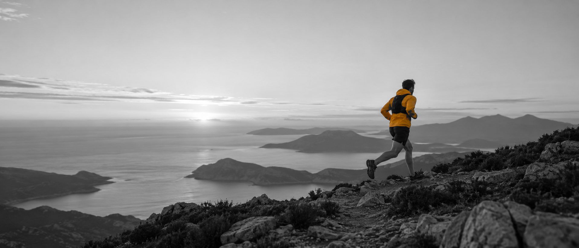 A trail runner in an orange jacket on a mountain path above the sea, placeholder photograph